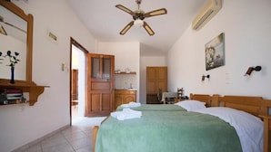 A bedroom with two beds, a ceiling fan, a kitchen in the background, and a door leading to another room. - Amphitrite Apartments (Kalymnos)