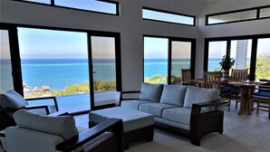 A spacious living room with a large sofa, a coffee table, and a dining area with wooden furniture. The room has large windows offering a view of the ocean and a lush landscape. - Stunning Turquoise views from this 2 bedroom, 2 bath home... (roatan)
