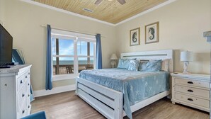 Ein Schlafzimmer mit Bett, Kommode, Nachttisch, Sessel, einem Fenster mit Vorhängen und einem Deckenventilator.