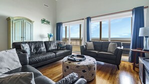 Ein geräumiges Wohnzimmer mit zwei schwarzen Ledersofas, einem runden Hocker mit nautischen Motiven, einem Holztisch und Blick auf das Meer durch große Fenster.