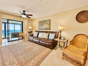 A living room with a brown leather sofa, a wicker chair, a glass coffee table, a floor lamp, and a painting on the wall.