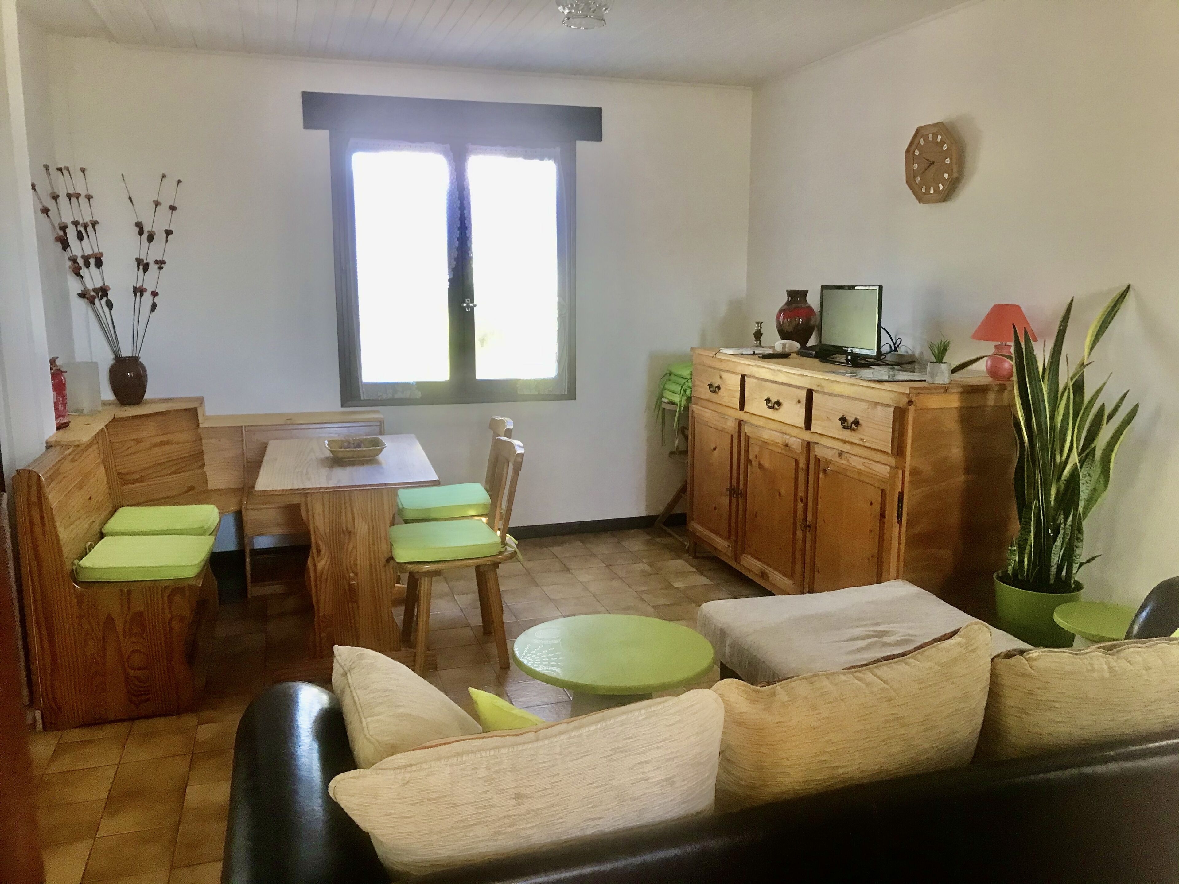 Een gezellige woonkamer met een bank, een eettafel met stoelen, een houten kast en een groot raam.