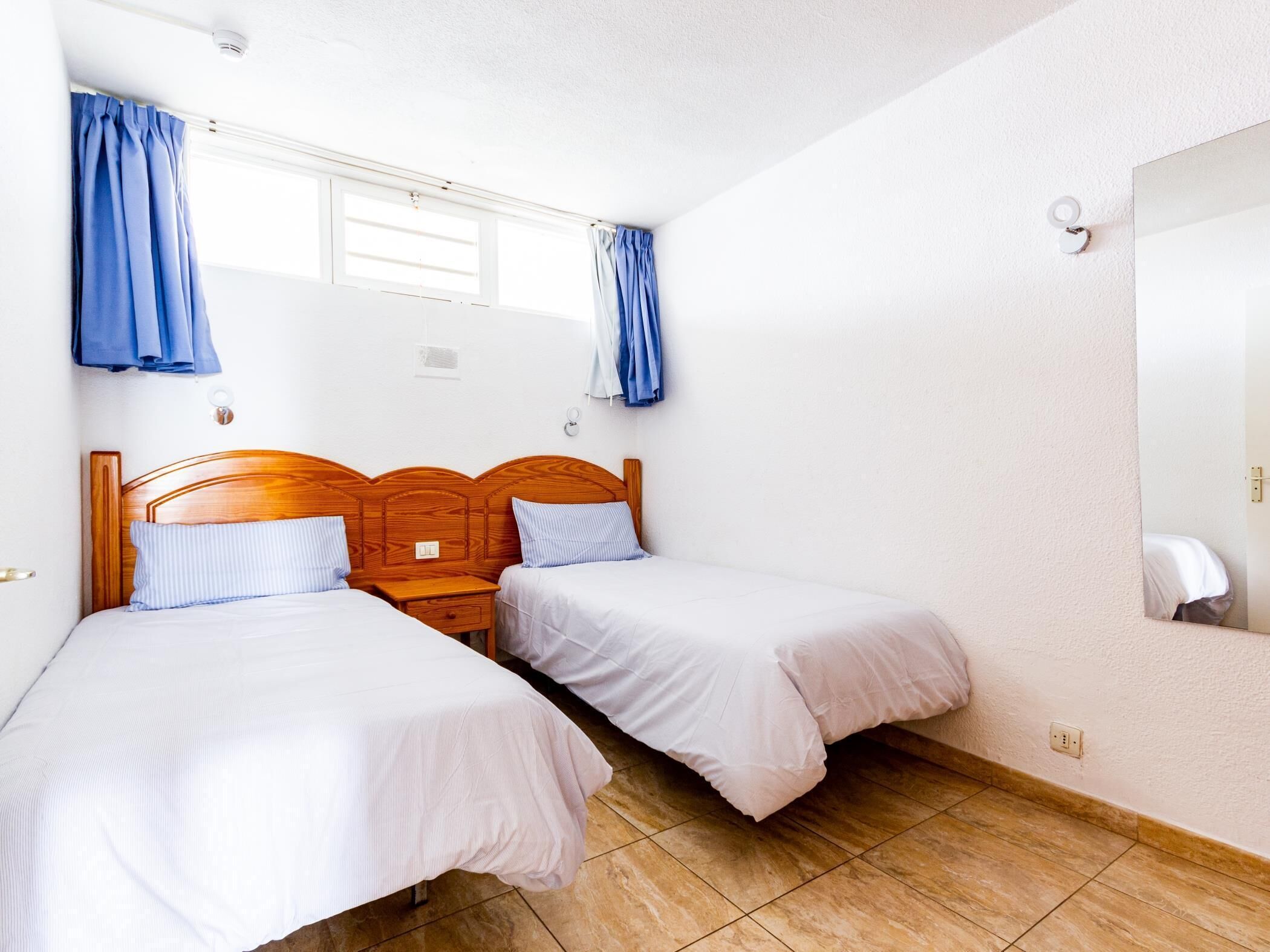 A hotel room with two single beds, a wooden headboard, a small bedside table, a mirror, and a window with blue curtains.