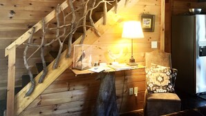 Eine Holztreppe mit einem verzweigtem Geländer, ein Schreibtisch mit Lampe, ein Stuhl und ein Kühlschrank.