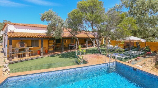 Una piscina con borde de azulejos, un patio con mobiliario exterior, y un edificio de techo y paredes amarillas.