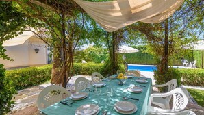 Zona pranzo all'aperto con tavolo apparecchiato per quattro, circondata da sedie bianche, sotto una tettoia.