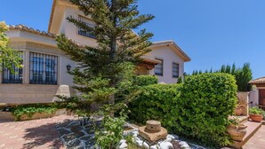 Un edificio in stile mediterraneo con tetto in tegole, vialetto in pietra e giardino curato con un albero grande e siepi.