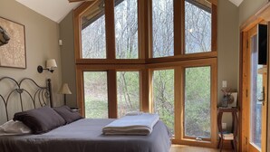 A bedroom with a large bed, wooden headboard, and a view of trees through large windows. - Irish Hill Chalet (West Valley)