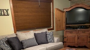 Una sala de estar con un sofá gris, cojines estampados, una mesa de centro de madera, un televisor sobre un mueble de madera y una ventana con persianas.