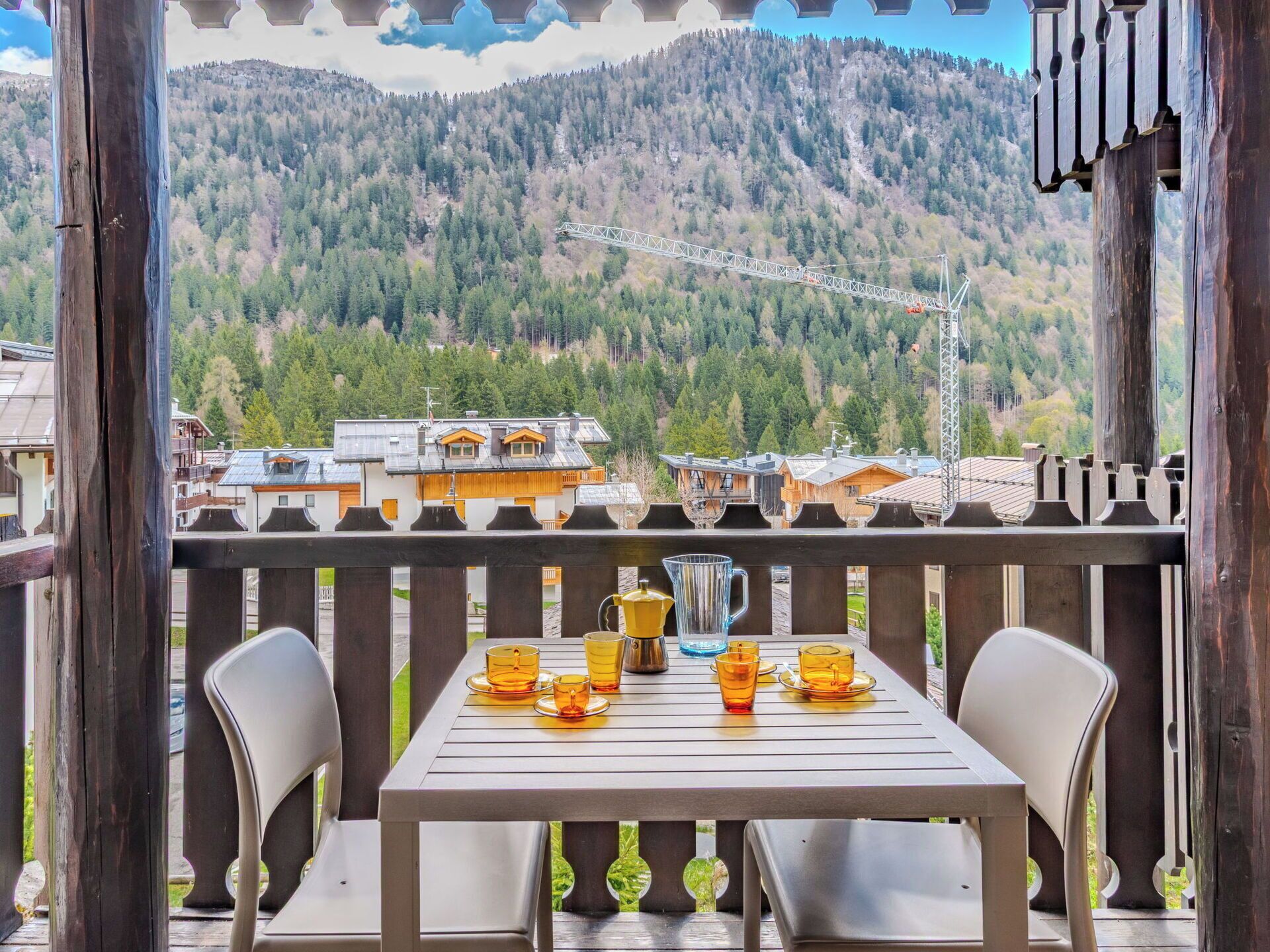 Terraza de madera con una mesa dispuesta para dos, con vistas a un paisaje montañoso y edificios.