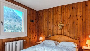 A bedroom with a wooden paneled wall, a bed with white bedding, a cross decoration, a window with a mountain view, and a radiator. - Lovely apartment for 5 guests with WIFI, TV, balcony and pets allowed (Ragoli)