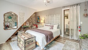 Un dormitorio con una cama, un baúl de madera, un muro de piedra y una cortina de macramé.