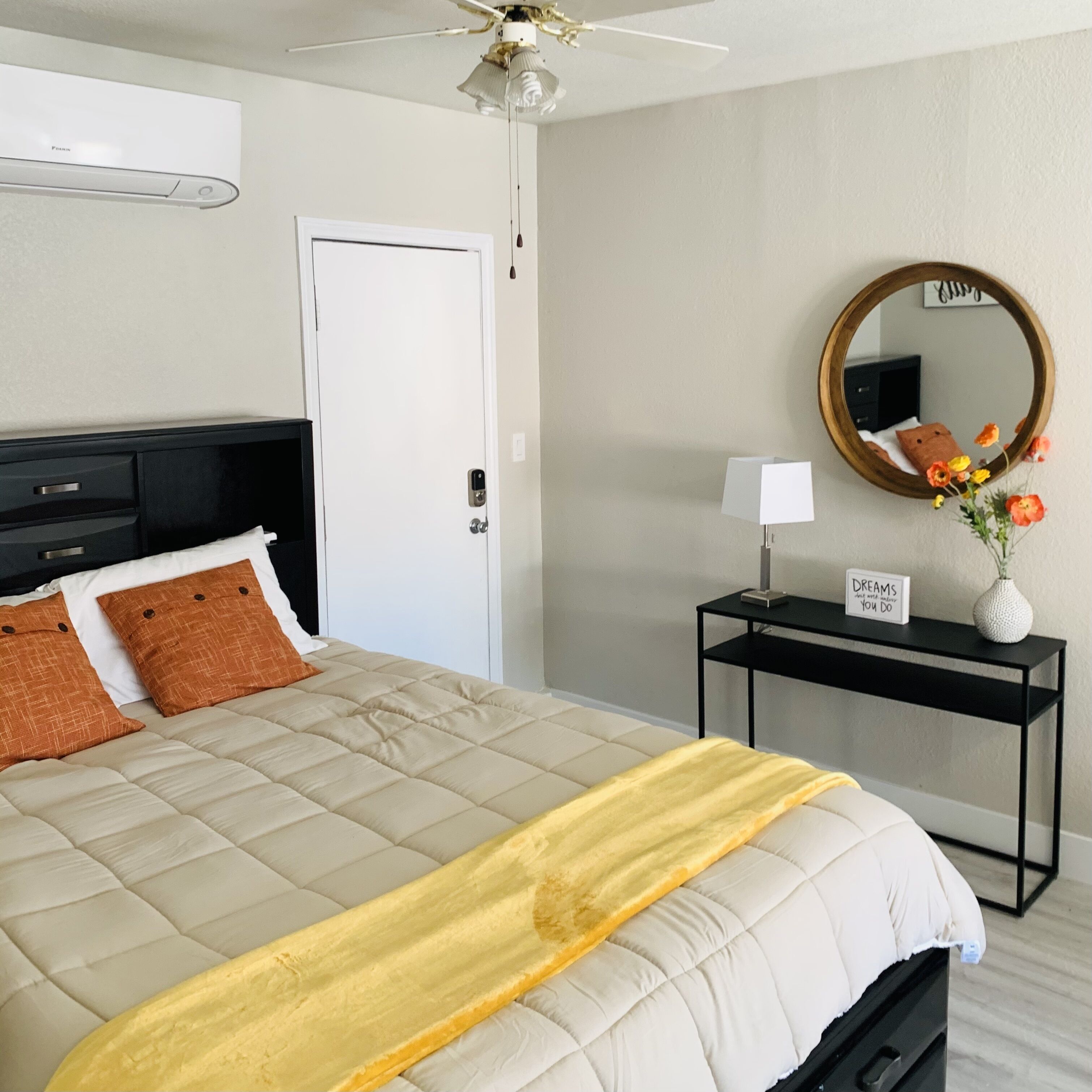 Quarto de hotel com uma cama, uma mesinha de cabeceira com uma luminária, um espelho e uma placa sobre a mesa.