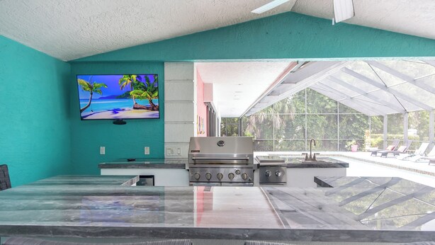 A kitchen with a large island, a TV mounted on the wall, and a view of the outdoor pool area. - Palm Palace - 2 House Compound Sleeps Up to 22!  Huge Pool and near the beaches (Fort Myers)