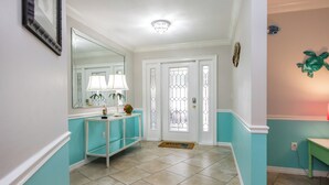 A hallway with teal walls, a white door with glass panels, a mirror, a teal console table with a lamp, and a turtle decoration on the wall. - Palm Palace - 2 House Compound Sleeps Up to 22!  Huge Pool and near the beaches (Fort Myers)