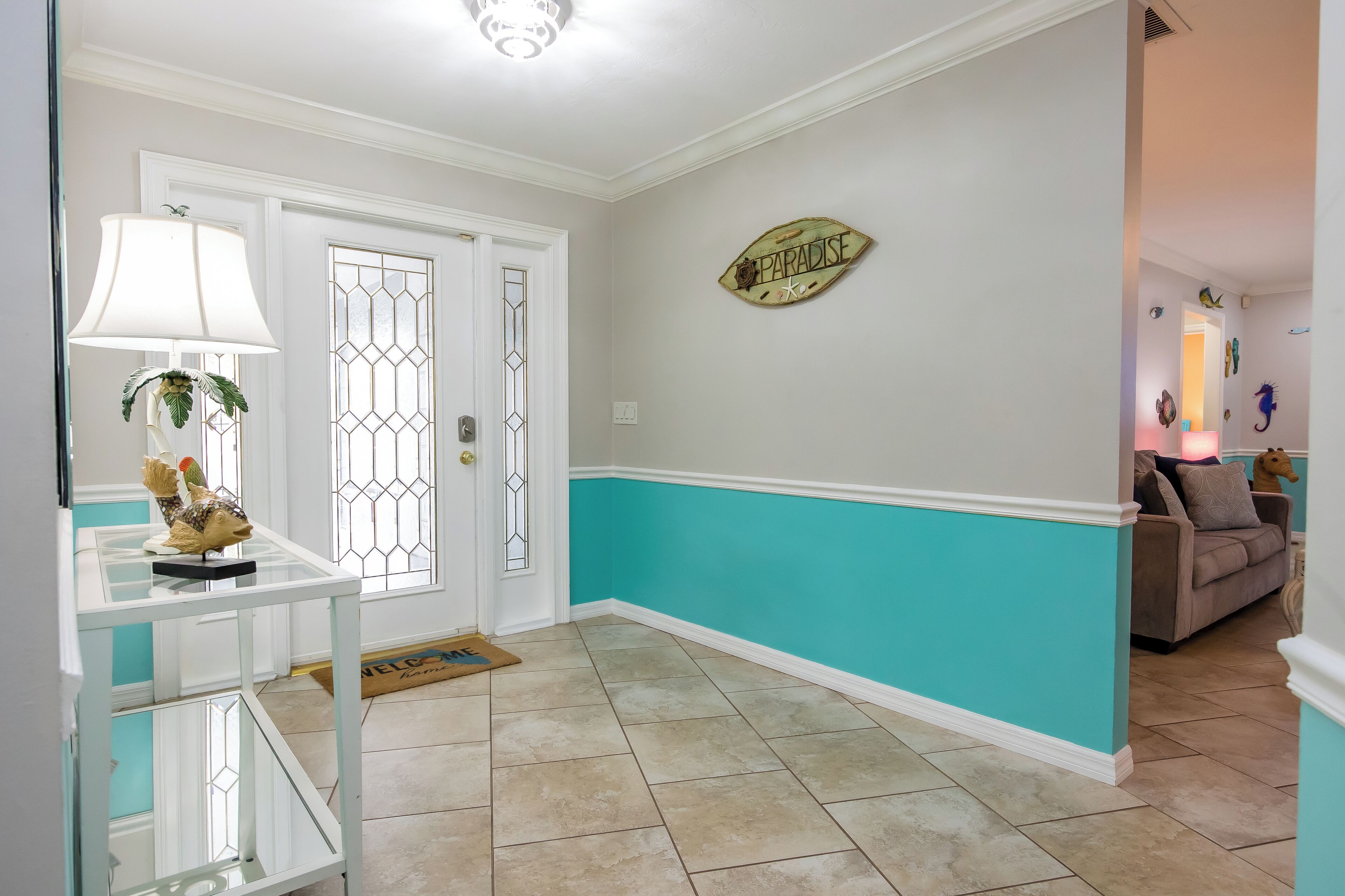 A hallway with a white door, a teal accent wall, a 'PARADISE' sign, and a brown sofa in the background.