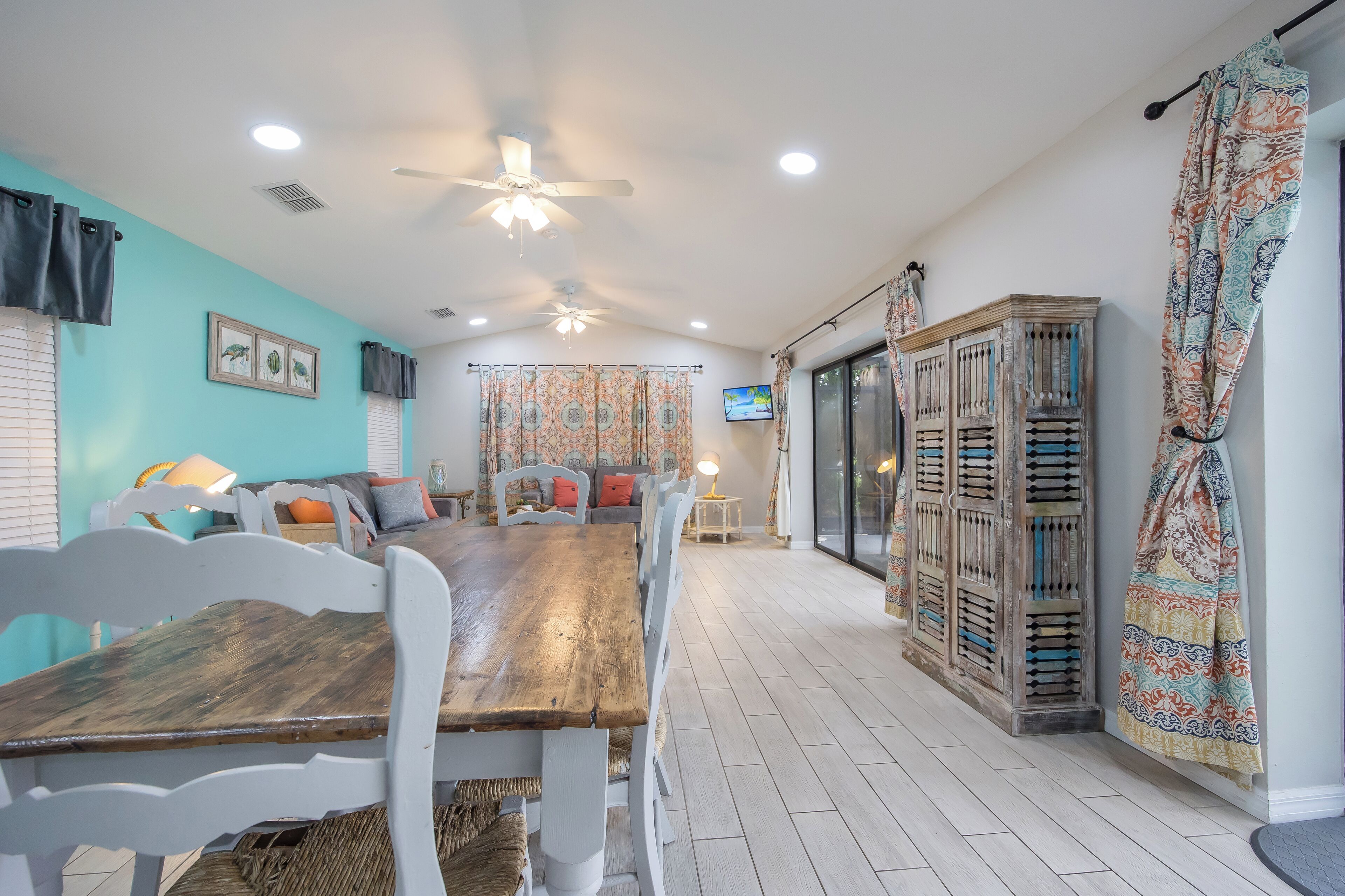 A dining area with a wooden table and white chairs, a ceiling fan, and a TV mounted on the wall.