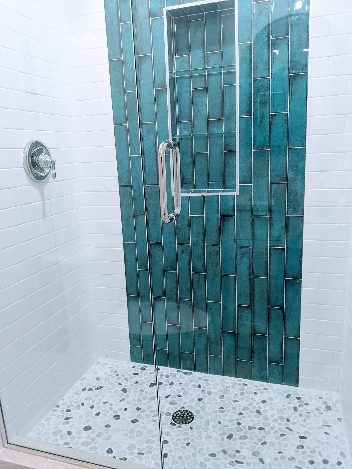 A shower area with a glass door, white tiled walls, and a pebble-patterned floor.