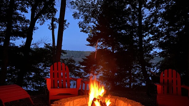 Ein Lagerfeuer, umgeben von Adirondack-Stühlen in einem bewaldeten Gebiet.