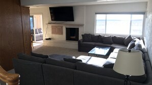 A living room with a fireplace, a large flat-screen TV mounted above it, a grey sectional sofa, and a large window offering a view of the sea. - ON THE SAND! (Oxnard)