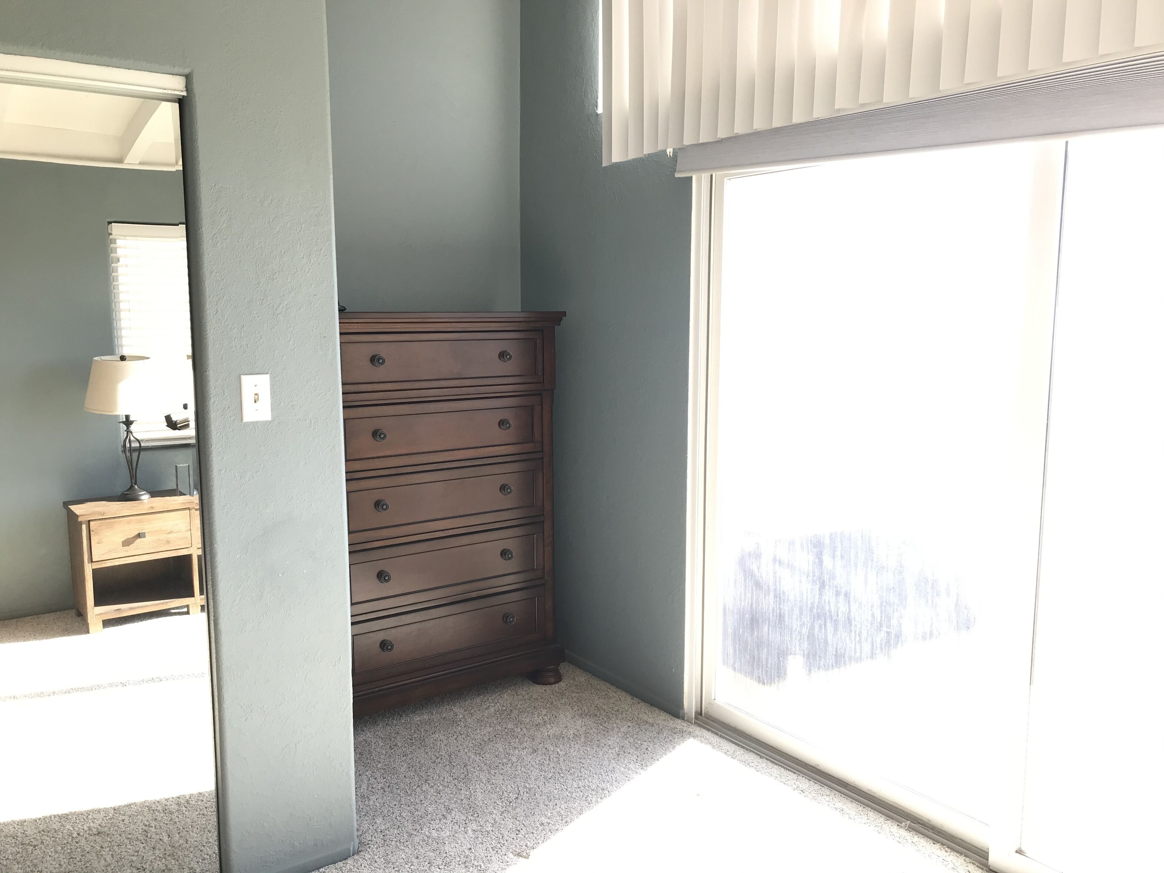 A room with a wooden dresser, a small table with a lamp, and a large window with blinds.