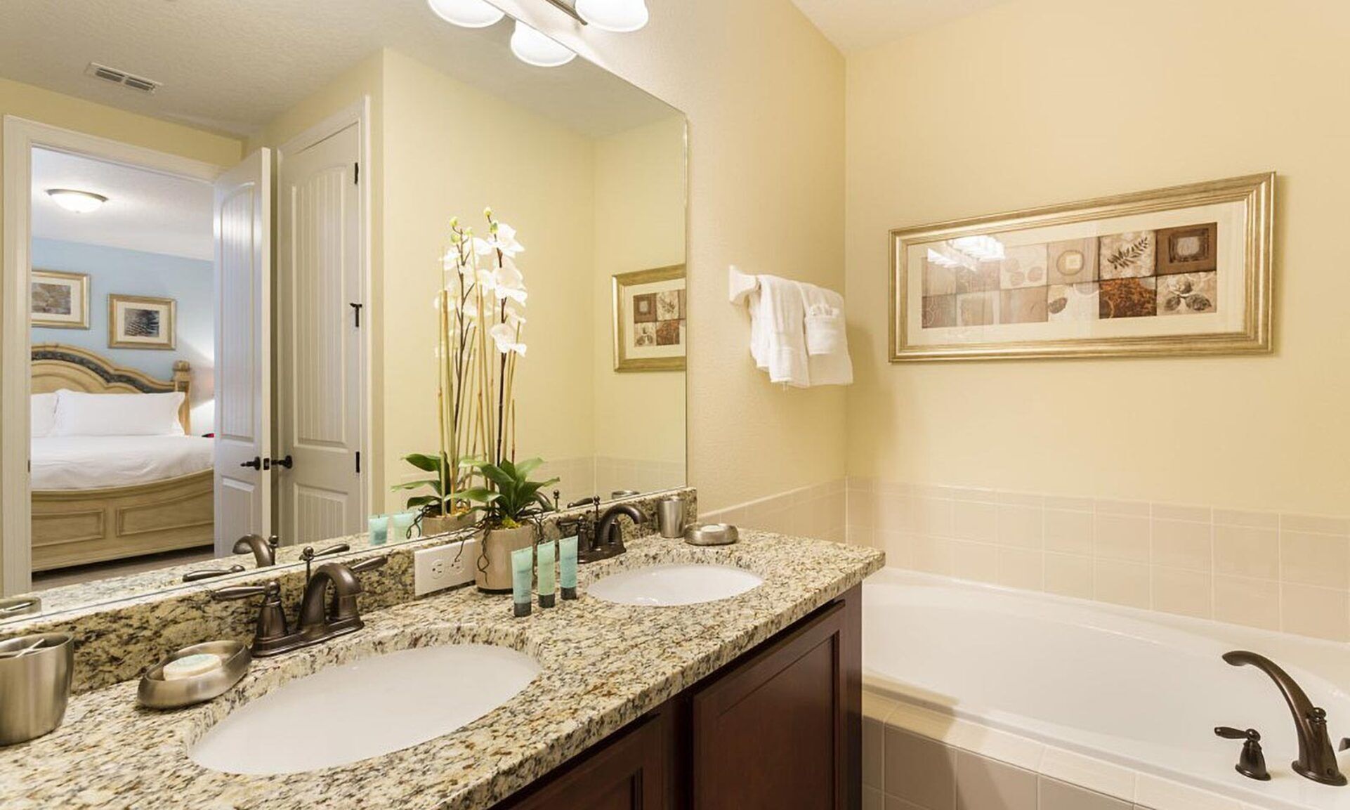 A hotel bathroom with two sinks, a large mirror, a bathtub, and a door leading to a bedroom.