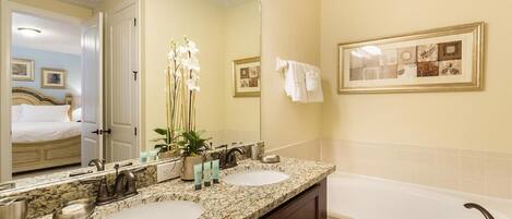 A hotel bathroom with two sinks, a large mirror, a bathtub, and a door leading to a bedroom.