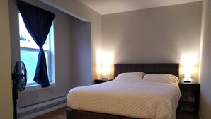 Een slaapkamer met een groot bed, twee bedlampen, een raam met gordijnen en een staande ventilator.