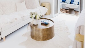Ein heller, weißer Innenraum mit einer weißen Couch, einem runden Couchtisch und Blick auf einen Außenbereich durch eine offene Tür.