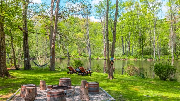Un espacio al aire libre tranquilo con un estanque, rodeado de árboles y con barriles de madera y una hamaca.