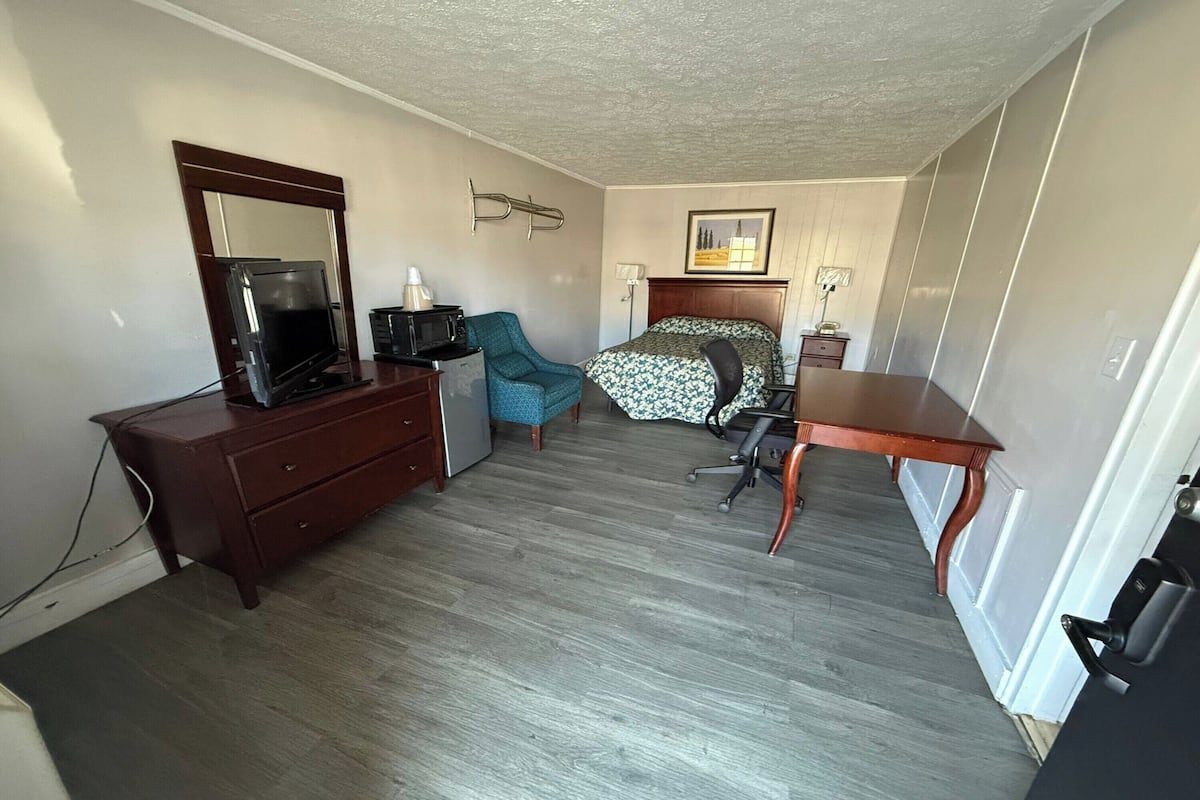 A compact hotel room with a bed, a desk, a chair, a dresser, a television, and a small table.