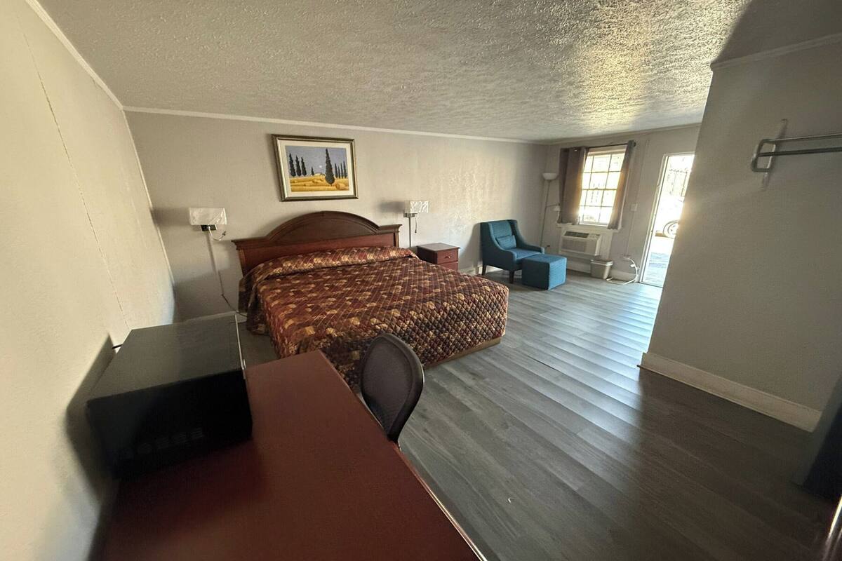 A hotel room with a bed, a desk, a chair, and a small table. There is a window with curtains and a painting on the wall.