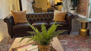 A living room with a dark brown leather sofa, two cushions, a woven basket with books, and a potted fern.