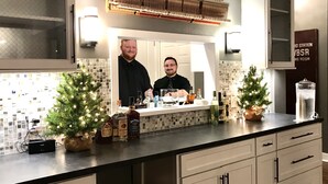 A kitchen with a piano, two potted Christmas trees, and a mirror reflecting two people.