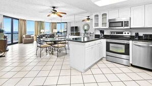 A modern kitchen with white cabinets, a central island, and stainless steel appliances. There is a dining area with a table and chairs, and a large window offering a view. - Beachfront 1BR condo in Destin with balcony, Gulf views and pool access (Destin)