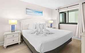 Een net opgemaakt bed met witte lakens en handdoeken in de vorm van harten, een nachtkastje met een lamp, een venster met gordijnen.