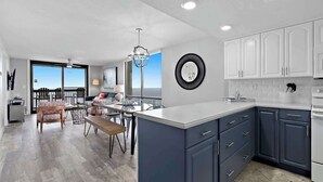 A modern kitchen with dark blue cabinets, a white island, and a dining area with a view. - SunDestin Resort Unit 1512 (Destin)
