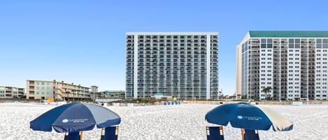 Una playa con dos sombrillas azules y sillones de playa, un gran edificio de hotel y otro edificio al fondo.