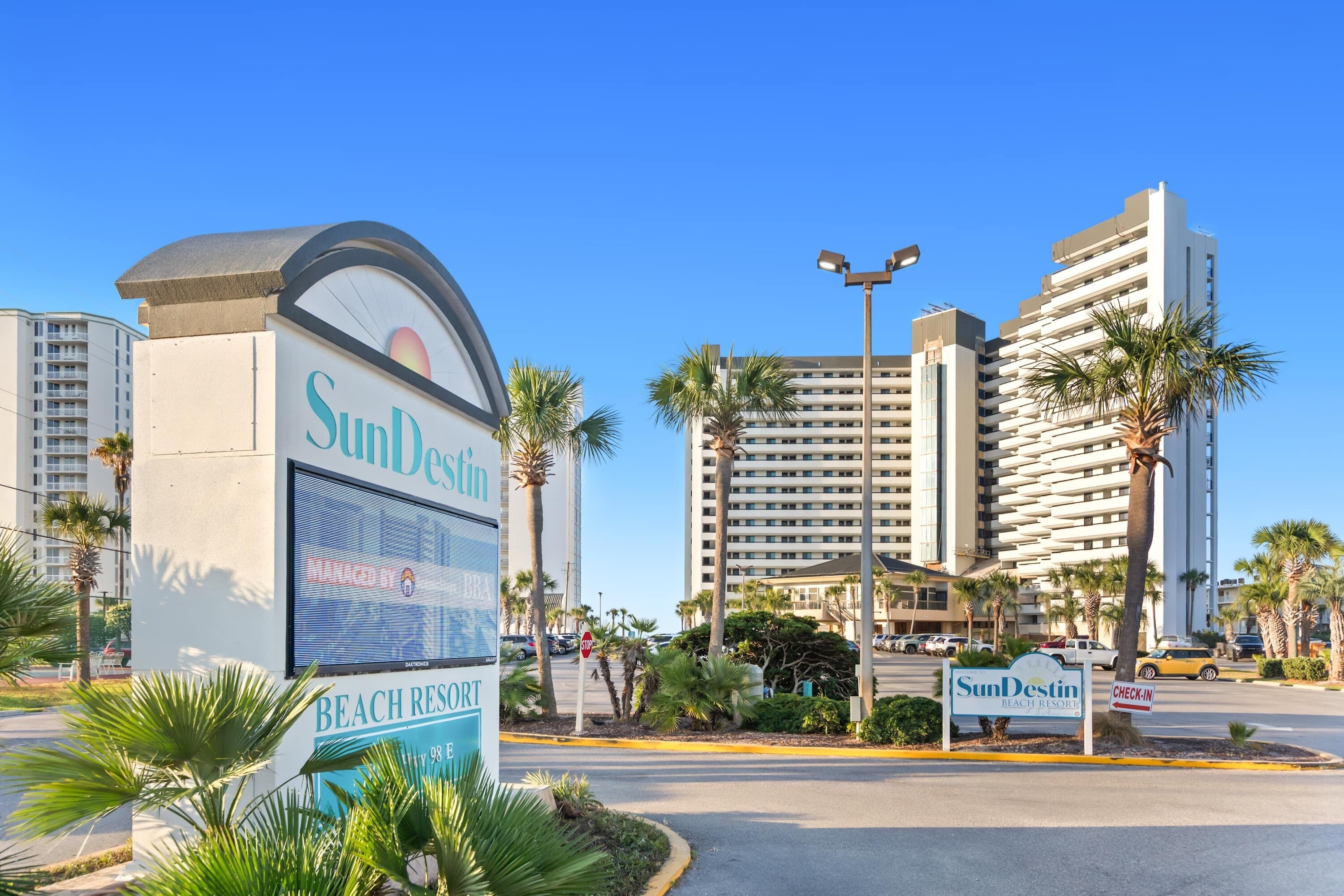 Een groot hotelschiltje met de tekst 'SunDestin Beach Resort' en 'Check-In', omringd door palmbomen en moderne gebouwen.