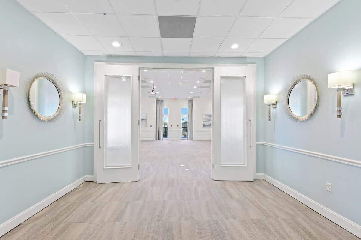 Un couloir d’hôtel aux murs bleu clair, aux moulures blanches et au plancher en bois clair. Au bout du couloir se trouvent deux portes, et des appliques murales ornées de miroirs ronds.