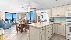 A spacious kitchen with a large island, wooden dining table, and chairs, with a view of the ocean through large windows. - SunDestin Resort Unit 1212 (Destin)