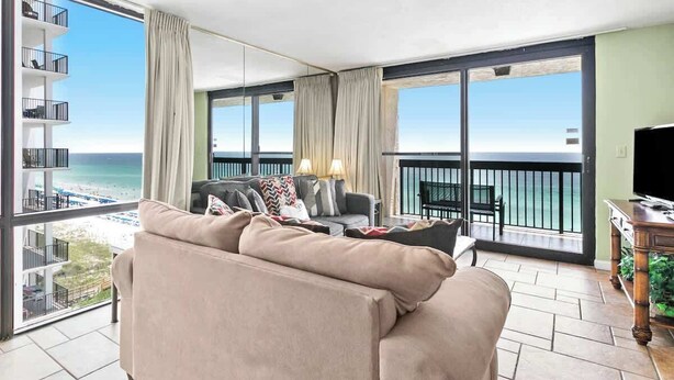Uma sala de estar ampla com uma grande janela com vista para uma praia, um sofá com almofadas, uma mesinha com abajures e uma TV em um móvel.