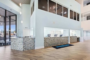 A modern hotel lobby with a reception desk, a second-floor balcony, and a view of the parking area.