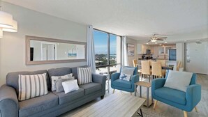 A modern living room with a grey sofa, blue armchairs, and a wooden coffee table. A large window offers a view of the city. - SunDestin Resort Unit 0712 (Destin)