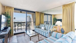 A modern living room with a sofa, a coffee table, and a TV. There is a balcony with a view of the city. - SunDestin 1BR Condo With Gulf-Front Sunrise and Sunset Views - Unit 0618 (Destin)
