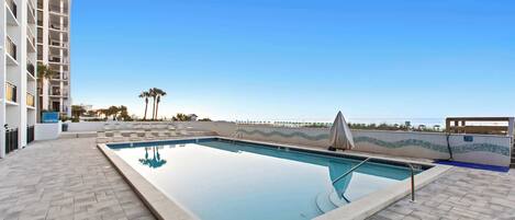 Una piscina en la azotea con agua azul cristalina, rodeada por una terraza con baldosas y un rascacielos al fondo.