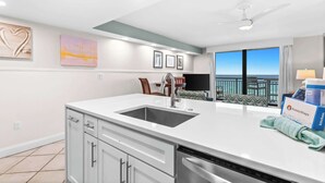 A modern kitchen with a large island, a dishwasher, and a view of the ocean. There are framed pictures on the wall and a ceiling fan.