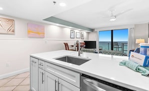 Una cucina moderna con una grande isola, lavastoviglie e vista sull'oceano. Si vedono quadri appesi alle pareti e una ventola a soffitto.