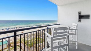 A balcony with white chairs and a table, overlooking a beach with umbrellas and people.