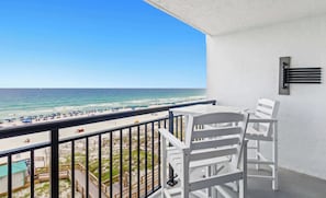 A balcony with white chairs and a table, overlooking a beach with umbrellas and people.